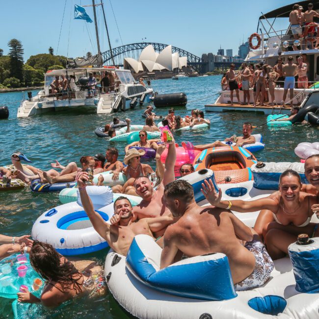 A vibrant scene of people enjoying a sunny day on a river, floating on inflatable rings and loungers. Boats are anchored nearby, and the Sydney Opera House and Harbour Bridge are visible in the background. The atmosphere is lively and festive.