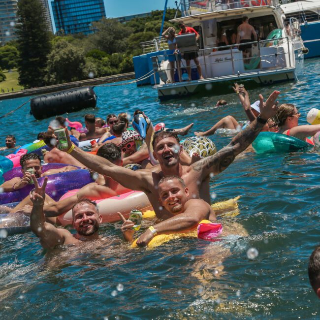 People are enjoying a sunny day in the water, surrounded by colorful inflatables. A boat is docked nearby, and several high-rise buildings can be seen in the background. Some individuals are posing playfully for the camera.