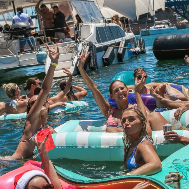 A group of people enjoying a sunny day on inflatable pool floats in the water, with clear blue skies and a bridge visible in the background. One person wears a Santa hat, adding a festive touch to the scene.