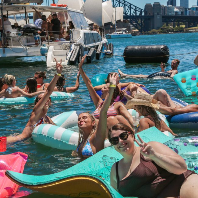 A group of people enjoying a sunny day on a river, floating on various colorful inflatables. A boat is anchored nearby, with more people visible onboard. The city skyline and a bridge are in the background.