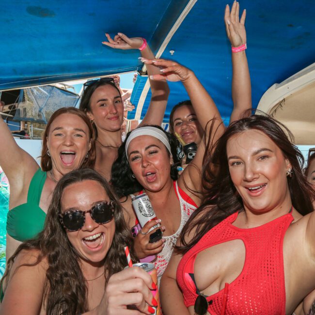 A group of women having fun on a boat, smiling and raising their arms in celebration. They appear to be enjoying a party atmosphere under a blue canopy.