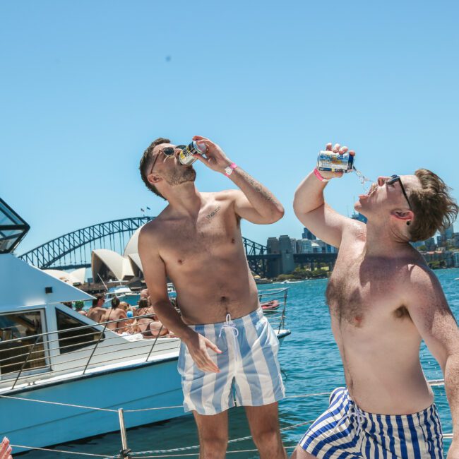 Two men in swim trunks stand on a boat, each drinking from a can. The background features a bridge and city skyline under a clear blue sky. Other people are visible on the boat, enjoying the sunny day.