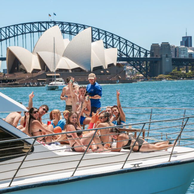 A group of people enjoying a sunny day on a boat in Sydney Harbour, with the Sydney Opera House and Harbour Bridge in the background. Several people are seated and standing, waving and smiling towards the camera.