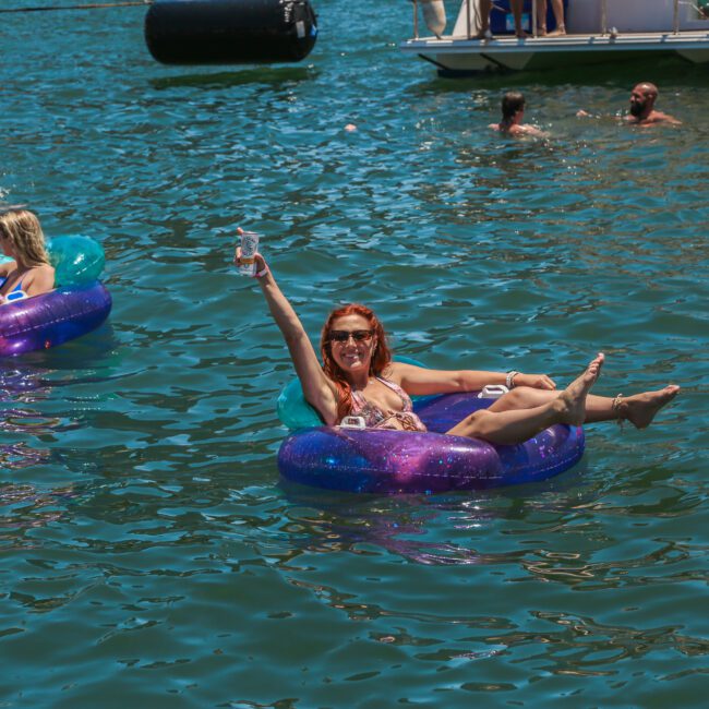 A woman in a swimsuit relaxes on a colorful inflatable ring in the water, raising a drink in celebration. Other people on floats and in the water are visible nearby. Sunny weather and a boat in the background complete the lively scene.