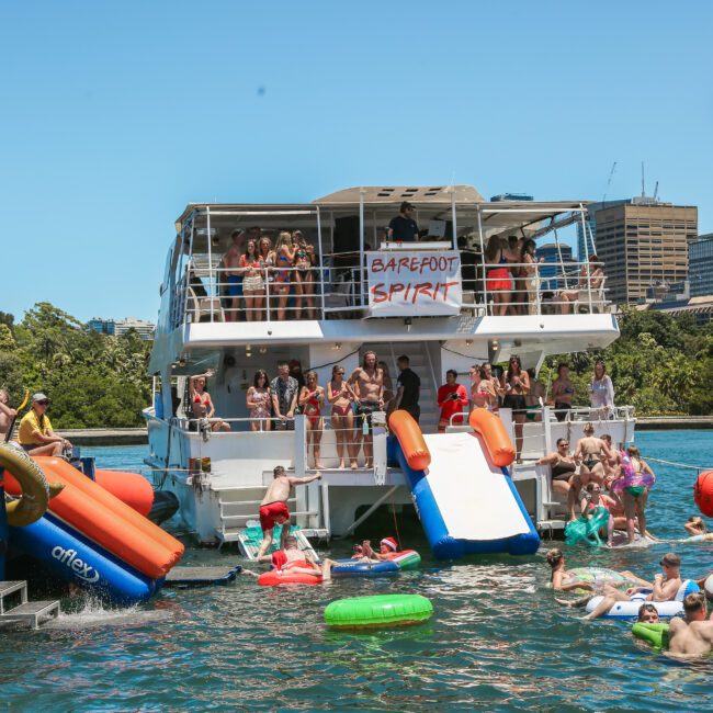 A large group of people enjoying a sunny day on a boat party. Many are on inflatables in the water, while others socialize on the boat's deck. A "Barefoot Spirit" banner is visible. The city skyline and lush greenery are in the background.