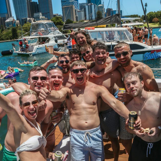 A group of smiling people in swimwear enjoying a sunny day on a boat in a harbor, with skyscrapers in the background. They are holding drinks and showing a joyful, celebratory mood.