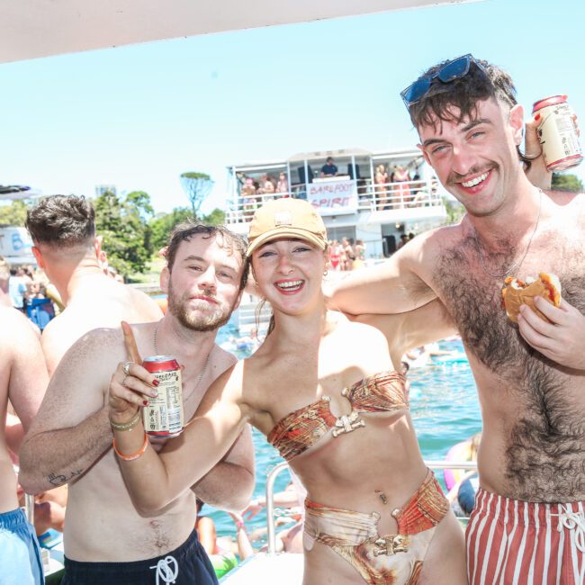 A group of smiling people enjoying a sunny day on a boat. One person holds a drink, and another has a sandwich. Other people are in the background, with clear skies above.