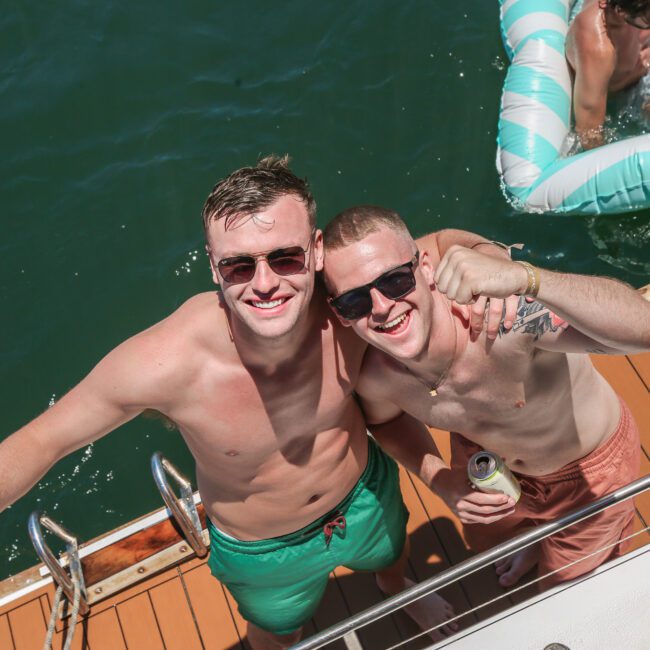 Two shirtless men in sunglasses smile at the camera on a boat. One holds a drink can, and an inflatable float with people is visible on the water nearby. The boat deck is wooden.