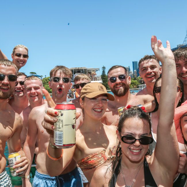 A group of people in swimwear smiles and raises drinks, enjoying a sunny day outdoors. Some wear sunglasses and hats. A city skyline and clear blue sky are visible in the background.