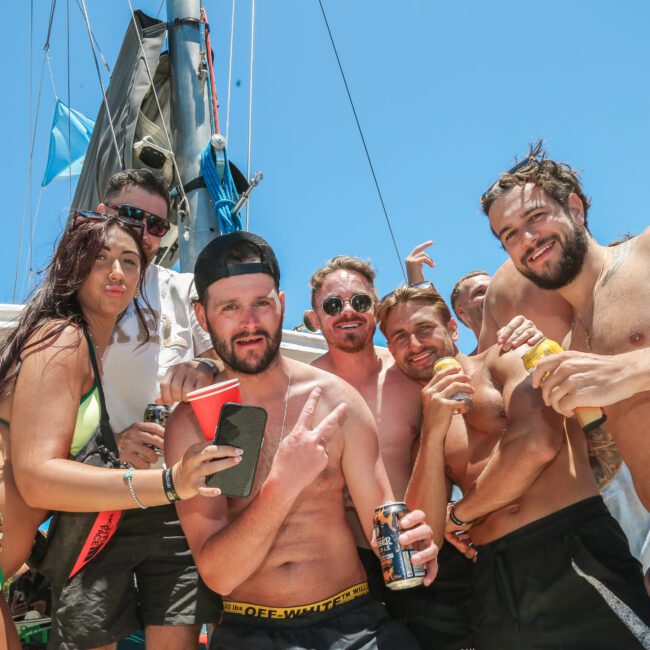 A group of people on a boat enjoying a sunny day. They are wearing swimsuits and holding drinks, smiling and posing for the camera. A mast and part of the sail are visible in the background.