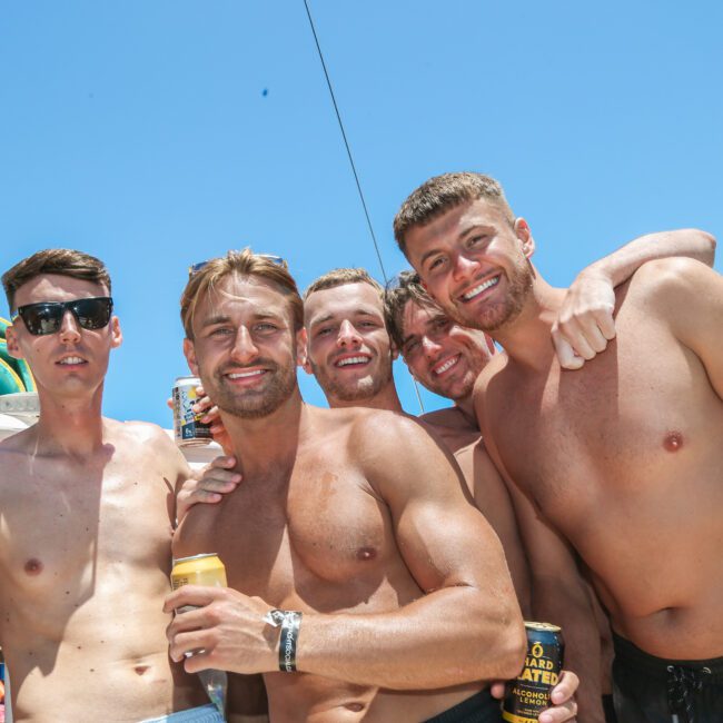 Five shirtless men are smiling and posing together on a boat under a clear blue sky. They are holding cans of drinks. One man is wearing sunglasses. A cityscape is faintly visible in the background.