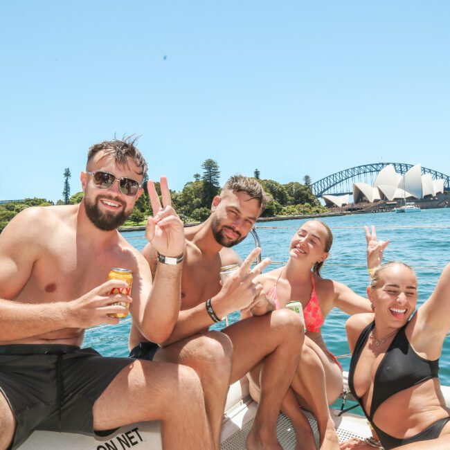 A group of four people in swimwear are sitting on a boat, smiling, and making peace signs. They are enjoying sunny weather with the Sydney Opera House and Harbour Bridge visible in the background. Two of them are holding beverage cans.