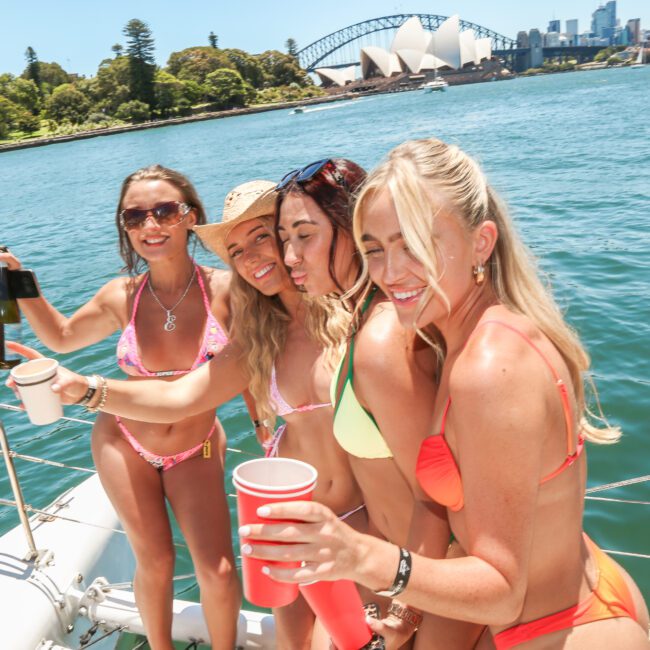 Four women on a boat in swimwear toast with red cups. They are smiling with sunny Sydney skyline, including the Opera House and Harbour Bridge, in the background.