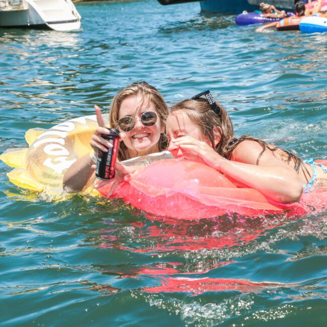 Two women smiling and holding drinks while floating on inflatable toys in a sunny body of water. One is wearing sunglasses, and they appear to be enjoying a summer day near boats.