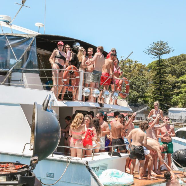 A group of people are gathered on and around a boat in swimwear, enjoying a sunny day on the water. Some are standing, and others are sitting. The background features trees and a clear blue sky.