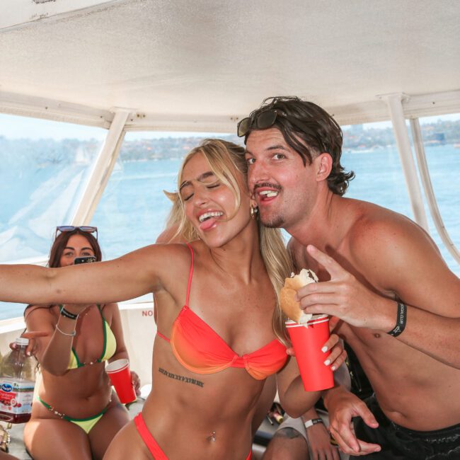 A group of people on a boat, wearing swimwear and holding red cups. One person is wearing an orange bikini, smiling with an arm around another person in sunglasses, shorts, and a hat. The background shows water and the horizon.