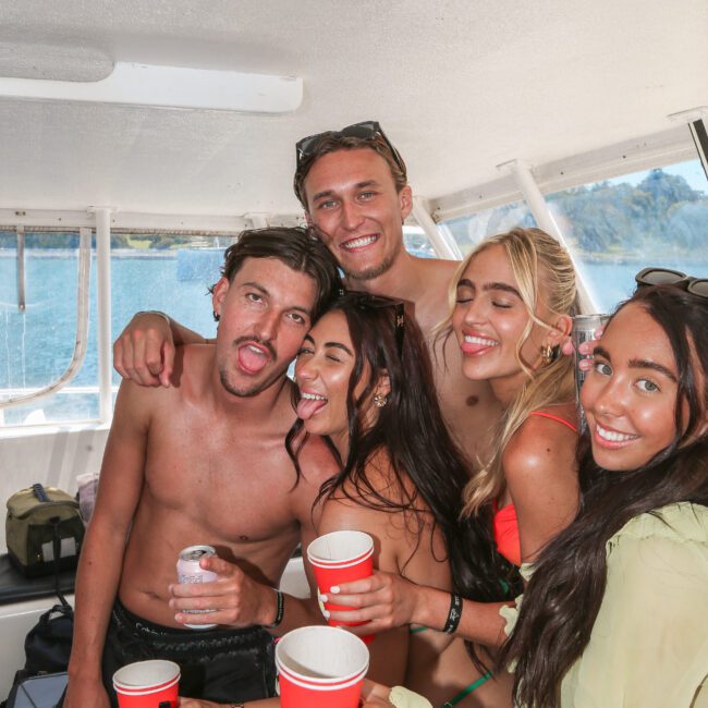 A group of people smiling and holding red cups on a boat. They are wearing swimwear and appear to be enjoying a sunny day near the water.