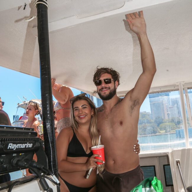 Man and woman in swimwear on a boat, with the man raising his arm and holding a red cup. They are surrounded by other people enjoying a sunny day on the water.