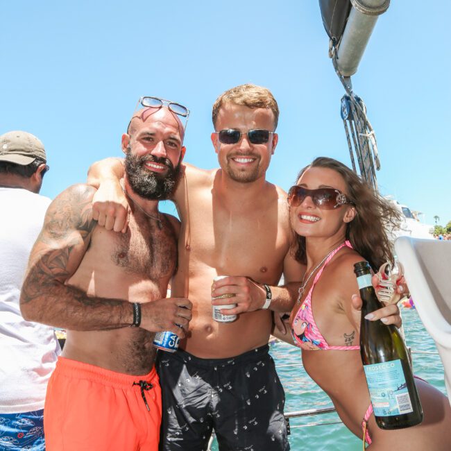 Three people smiling on a boat under clear blue skies. Two men and a woman in swimwear are holding drinks. The man on the left has a beard and sunglasses, while the woman is holding a bottle. They appear to be enjoying a sunny day at sea.