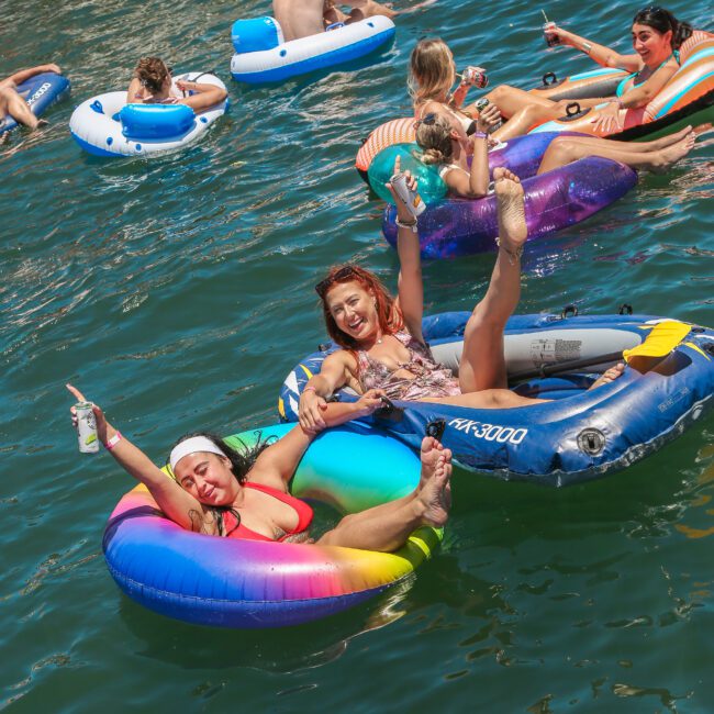 A group of people are enjoying a sunny day on a lake, floating on inflatable rafts in vibrant colors. Two women in the foreground are smiling and holding drinks, relaxing on a rainbow-patterned tube.