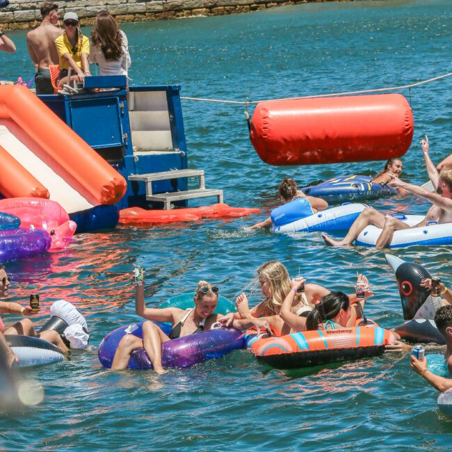 People are enjoying a sunny day at the lake, lounging on colorful inflatables. There are floating structures and a DJ booth nearby. The atmosphere is lively and festive, with attendees relaxing and socializing in swimsuits.