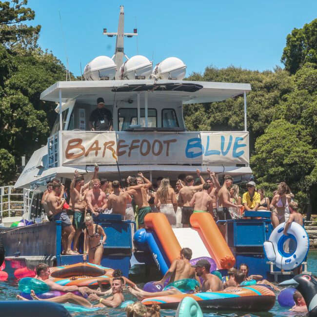 A large group of people are having a lively party on a boat named "Barefoot Blue." The scene includes inflatable floaties in the water and a waterslide coming off the back of the boat. Trees and clear blue skies are visible in the background.