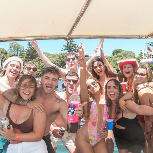 A group of people in swimwear smiling and posing together on a boat under a canopy. They are holding drinks and making celebratory gestures. Another boat and tree-covered hills are visible in the background, suggesting a lively outdoor gathering.