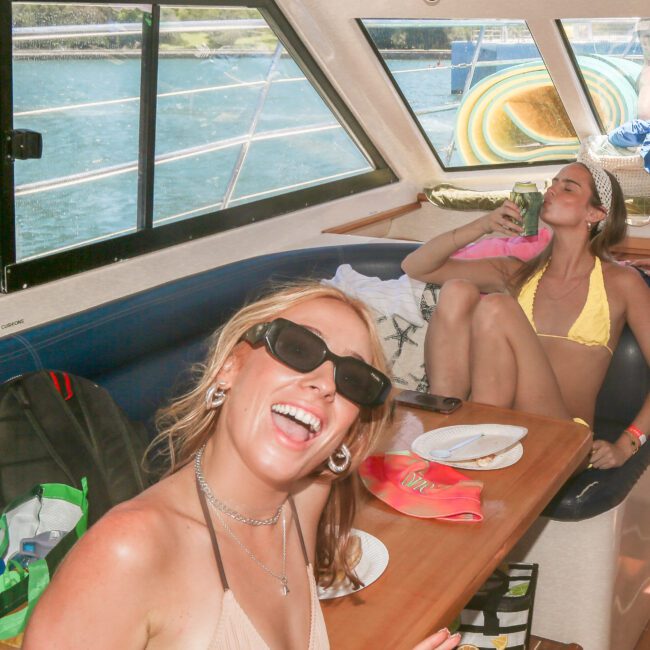 Two women in swimsuits relax inside a boat. One sits on a bench, smiling and posing for the camera; another lounges in the background, resting with closed eyes. A window shows a view of the water outside. Various items are scattered on the table.