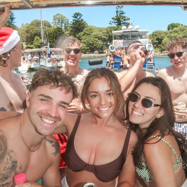 A group of young adults enjoying a sunny day on a boat. They're wearing swimsuits, holding drinks, and smiling at the camera. A man in a Santa hat is in the background, along with trees and a body of water. It’s a lively, festive atmosphere.