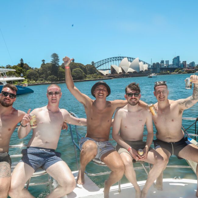 Five people pose while sitting on a boat in sunny weather, each holding a drink. The Sydney Opera House and Harbour Bridge are visible in the background, surrounded by water and city skyline.