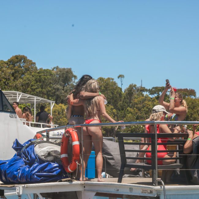 A group of people enjoying a sunny day on a boat. Some are wearing swimwear and embracing. Others are taking photos. The scene captures a festive atmosphere with clear skies and trees in the background.