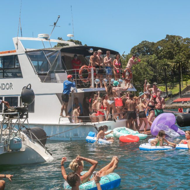 A lively scene with people on a boat and floating in the water, enjoying a sunny day. The boat is crowded, and several colorful inflatables are in the water. Trees and greenery are visible in the background.