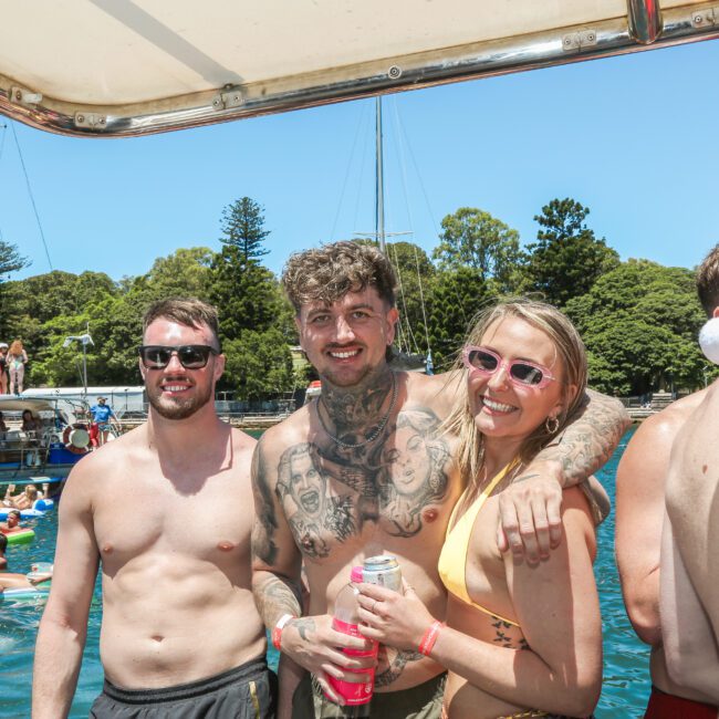 A group of people enjoying a sunny day on a boat. Three people are smiling at the camera; one has tattoos, and another is wearing a Santa hat. Other boats and people are in the background, with trees on the shore. The scene is lively and festive.