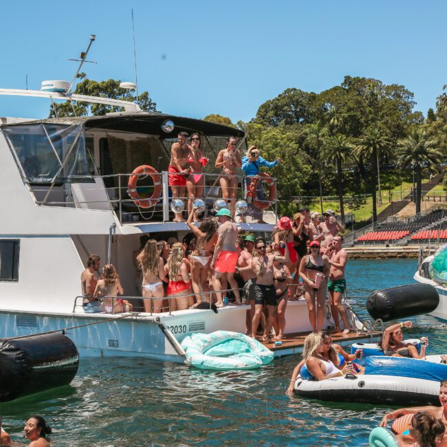 A lively scene on a sunny day with a group of people enjoying a party on a boat docked near the shore. People are swimming in the water, lounging on inflatables, and socializing on the boat deck. Trees and a park are visible in the background.