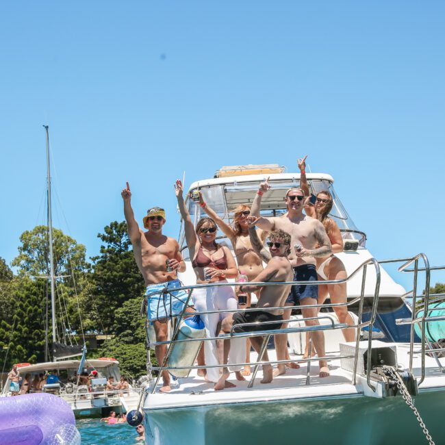 A group of six people, including adults and children, are standing on the deck of a boat, smiling and pointing up. They are wearing swimwear, with clear blue skies and trees visible in the background. Inflatable tubes are on the boat.