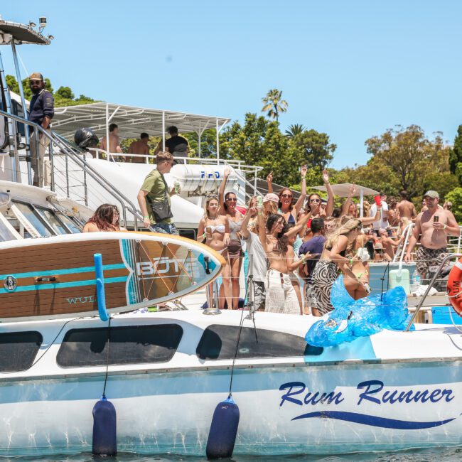 A group of people enjoying a sunny day on a boat named "Rum Runner." Some are sunbathing, while others are preparing to jump into the water. A paddleboard is visible, and trees line the background.