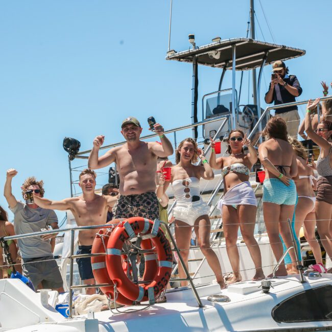 A group of people enjoying a sunny day on a boat, with several individuals holding drinks. They appear to be celebrating or having a party. The boat's deck is lively, and there's a clear blue sky in the background.