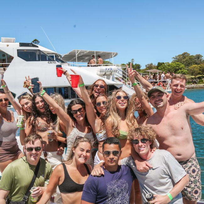 A group of people smiling and posing on a boat during a sunny day. Some hold drinks, and they're surrounded by clear water and trees in the background. The mood is festive and relaxed.