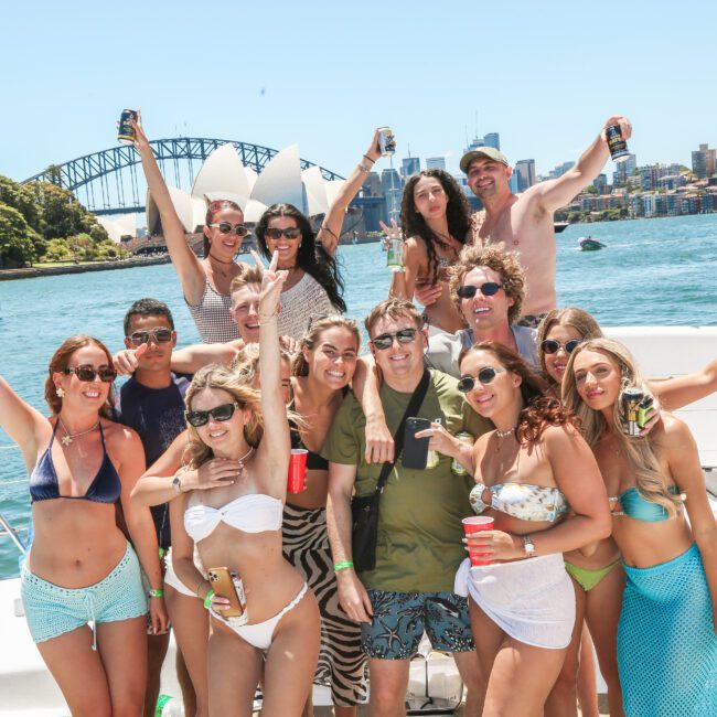 A group of people enjoying a sunny day on a boat. They are smiling and posing with drinks. The background features a bridge and skyline. The atmosphere is festive and relaxed.