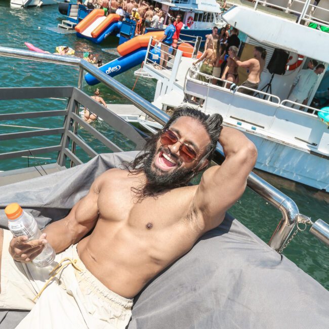 A shirtless man with long hair and sunglasses relaxes on a boat, holding a bottle. In the background, people enjoy a floating water park and slides near several boats on a sunny day.