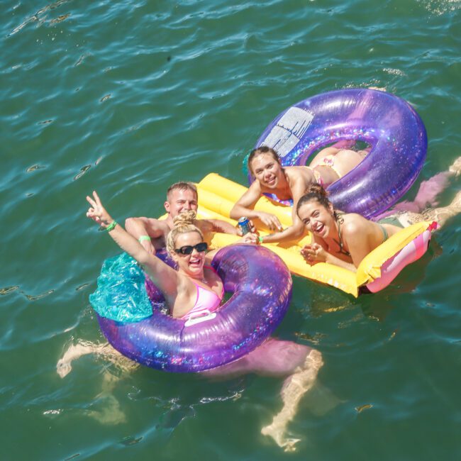 Four people smiling and relaxing on inflatable floats in a body of water. Two have purple ring floats, and the others are on a yellow float. The water is a deep green, and they appear to be enjoying a sunny day. One person is making a peace sign.