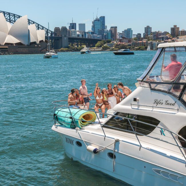 A group of people enjoy a sunny day on a boat in Sydney Harbour, with the Sydney Opera House and Harbour Bridge in the background. The boat has a rolled-up mat on the deck and clear blue water surrounds the scene.