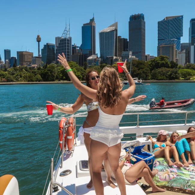 Two women in swimwear dance with red cups on a boat in a sunny harbor. Several people relax nearby on the deck. The city skyline and lush greenery are visible in the background under a clear blue sky.