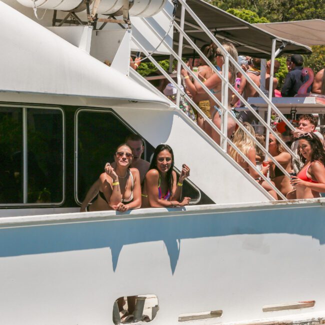 A group of people enjoying a sunny day on a boat. Two friends lean out of a window, smiling. The deck above is crowded with people in swimwear, sunbathing and socializing. The boat is surrounded by lush green trees.