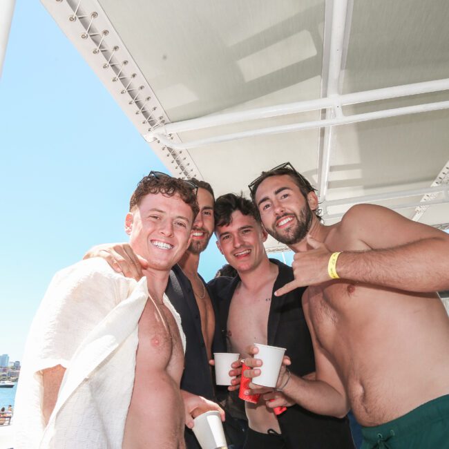 Four men are smiling and posing together on a boat under a canopy. Two are shirtless, holding drinks, and the day is sunny with a clear blue sky. The background shows a city skyline and water.