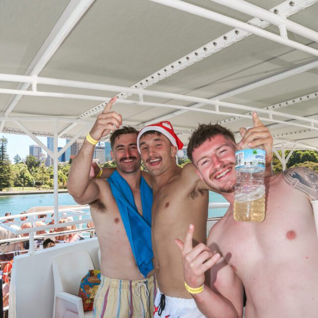 Three smiling men in swimwear stand on a boat, with one holding a water bottle. They are making celebratory gestures, surrounded by a sunny outdoor setting and urban skyline in the background.