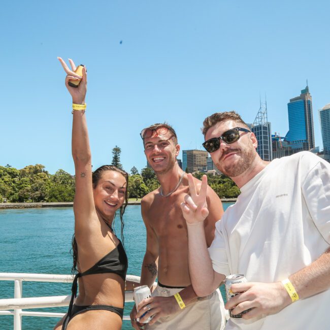 Three people smiling and posing for the camera on a boat with a city skyline in the background. Two are making peace signs, and one has their arm raised. It's a sunny day, and they are wearing casual summer clothes.