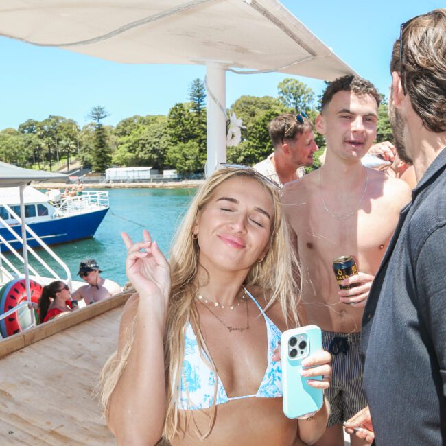 A woman in a blue and white bikini poses with a peace sign and closed eyes on a boat. She holds a phone in one hand. Other people, including a shirtless man, are in the background. The boat is on water, and the sky is clear.