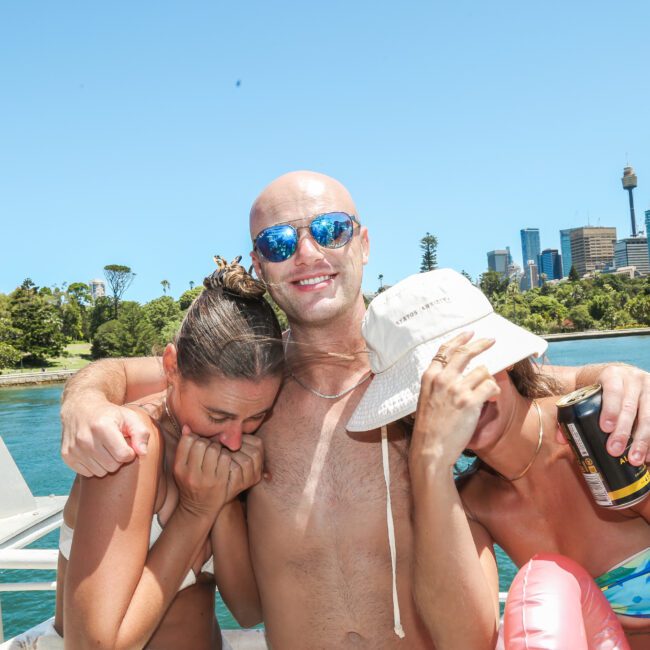 Three people are smiling on a boat with a city skyline in the background. The man in the middle is shirtless and wearing sunglasses. The two women beside him are leaning on his shoulders. One is holding a drink can. It's a sunny day.