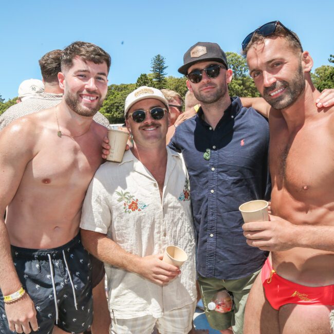 Four men posing on a boat under a clear blue sky. They appear relaxed and are dressed in casual summer clothing, some wearing swim shorts and sunglasses. A nearby ferry and green trees are visible in the background.